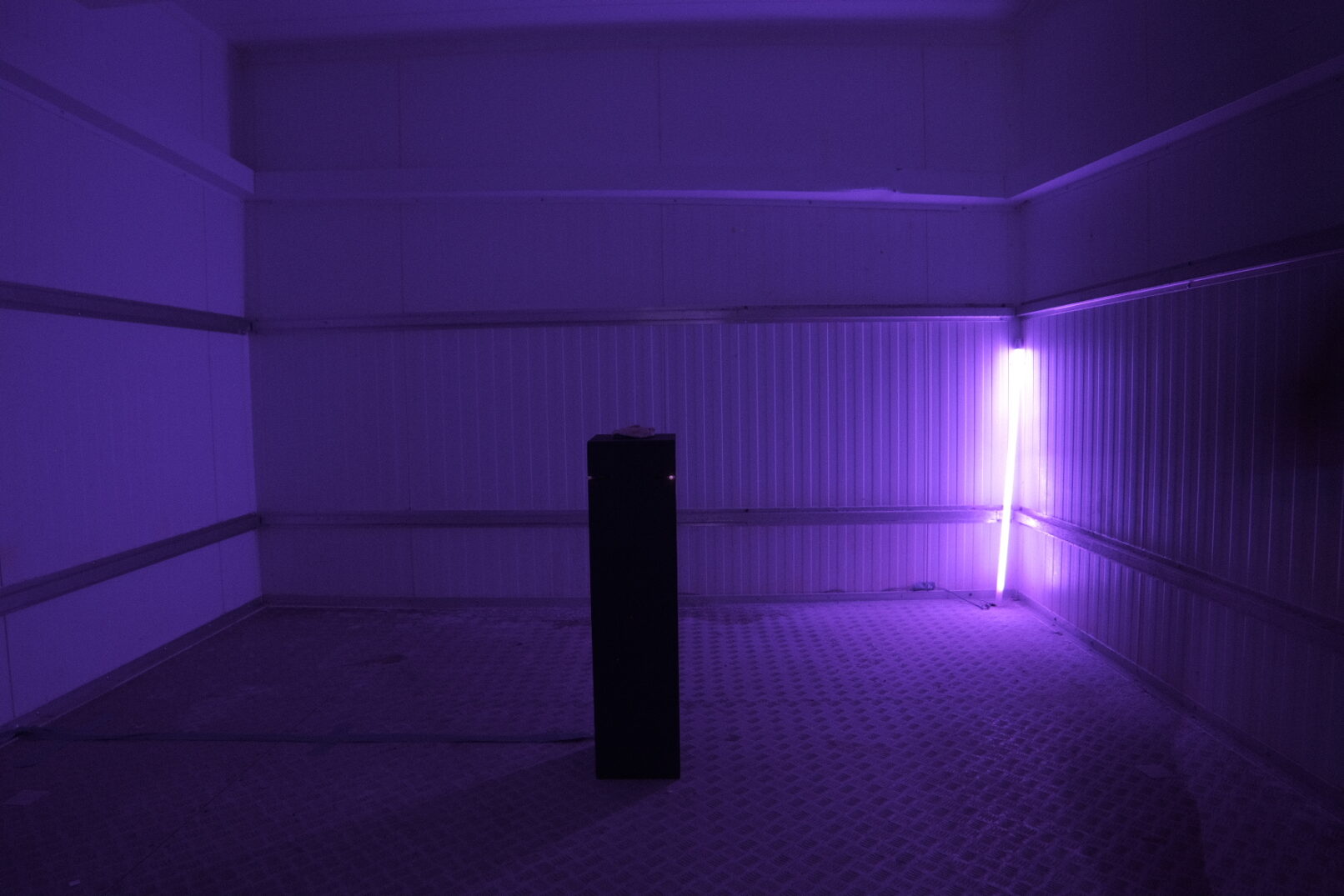 The installation "look for yourself or a human rights graveyard?" by Stathis Doganis. A small sculpture in a plinth made in the island of Lesvos. A lamp with purple light behind the sculpture is dimmed down when the visitor approaches the artwork.